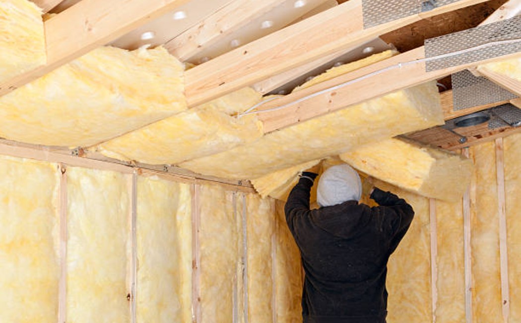 Worker installing pink fiberglass batt insulation between wooden trusses