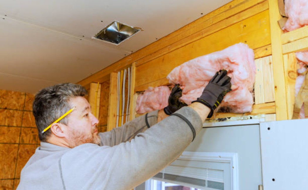 Worker placing fiberglass insulation between wooden studs in a house.